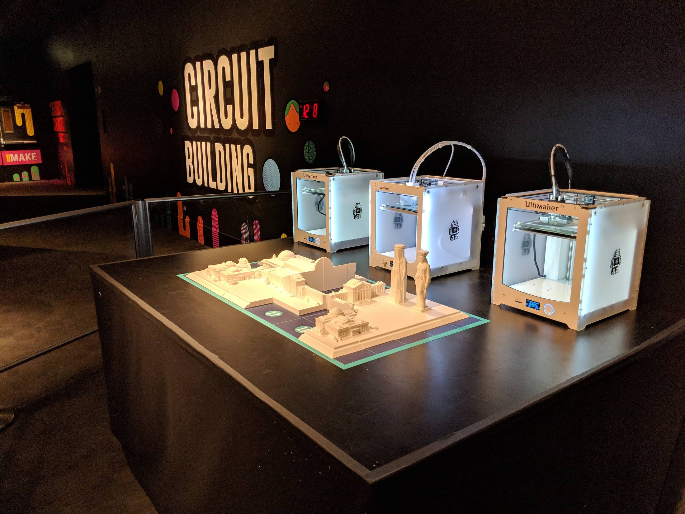 Wide view of the exhibit table with three Ultimaker printers and the growing white model in front of a dark wall lettered Circuit Building.