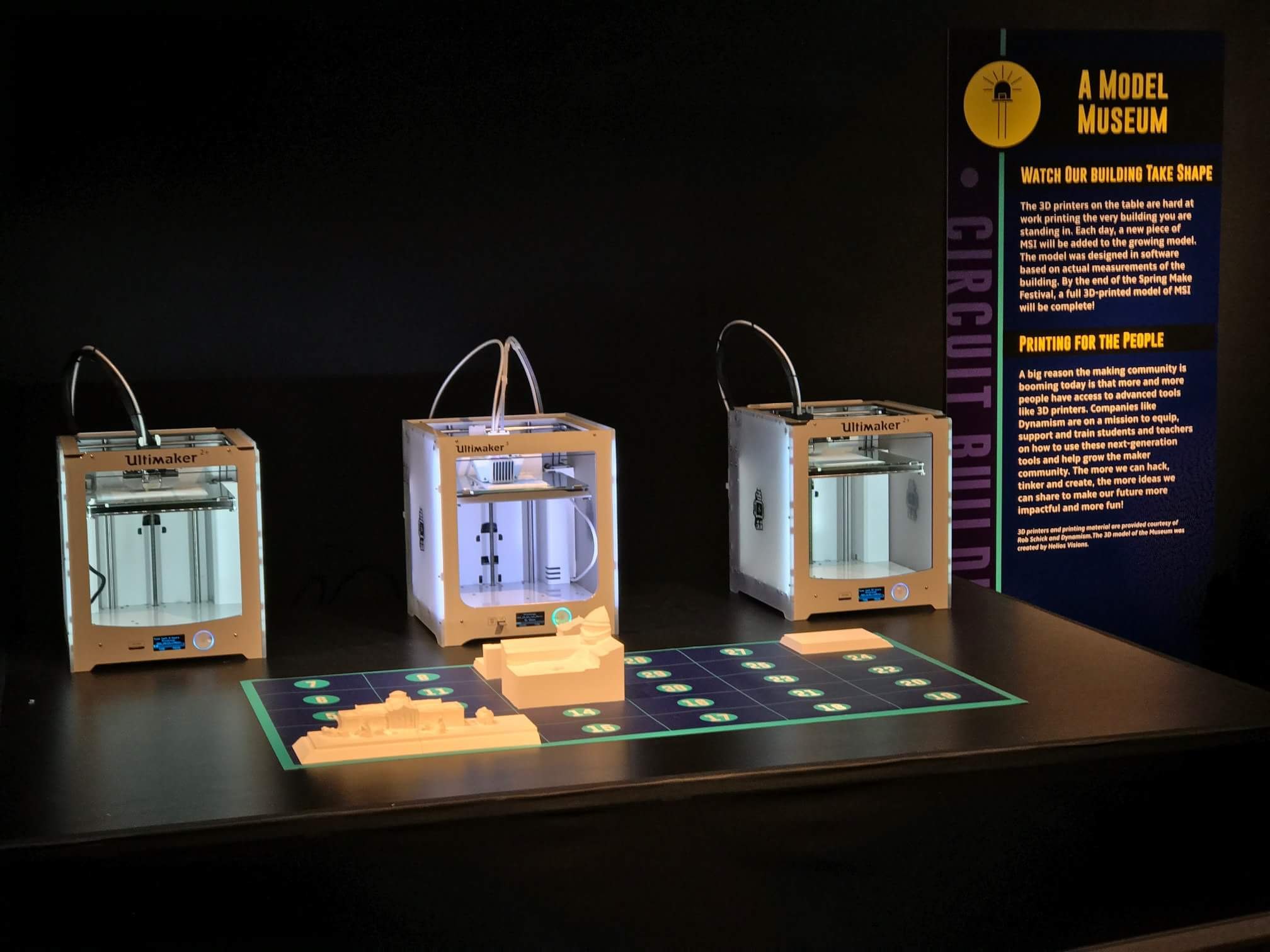 Three Ultimaker printers on a dark exhibit table behind a partly built white model, with empty grid squares still visible.