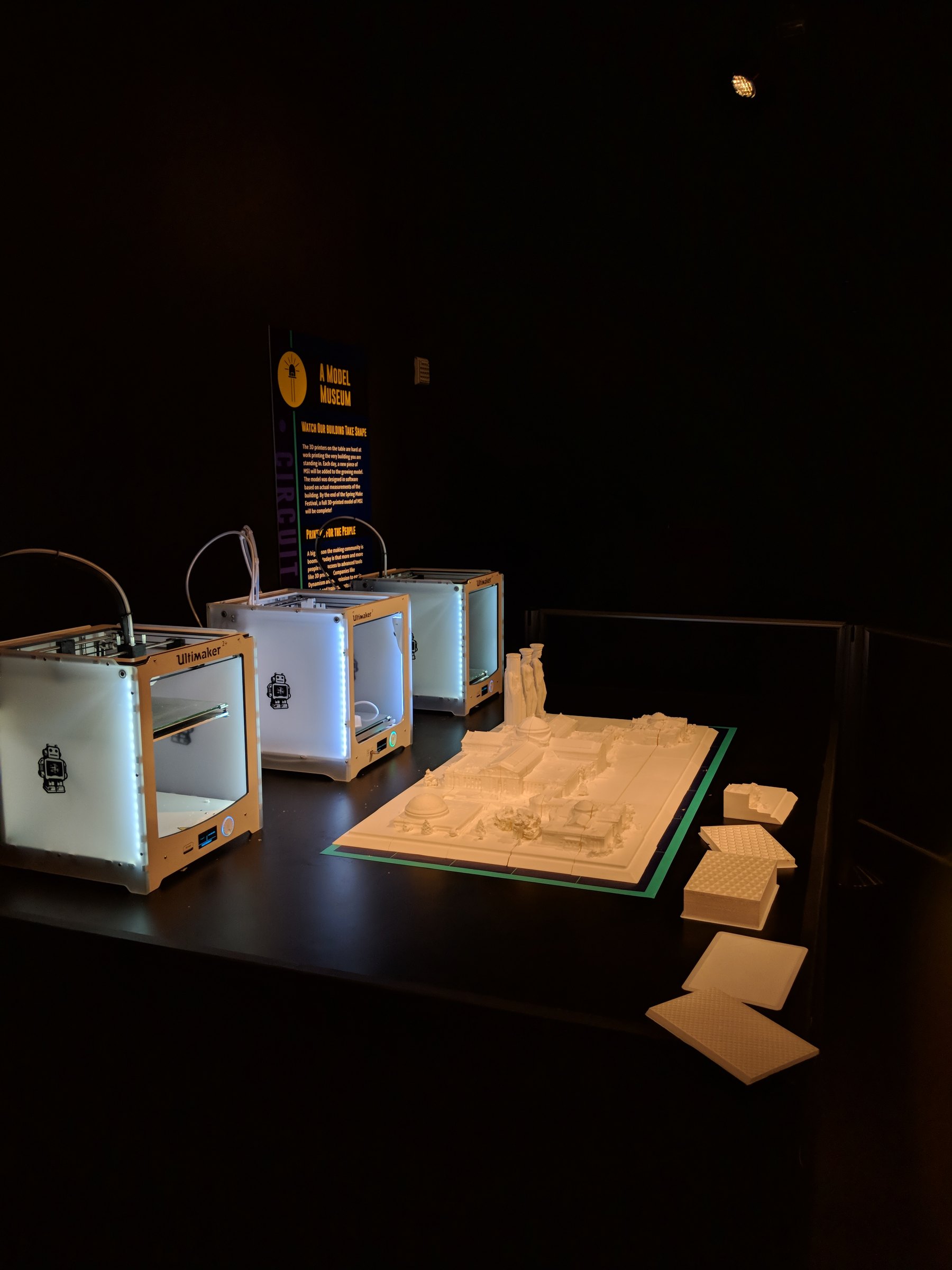 The exhibit table in low light on the festival's final day, with the assembled model, three printers and a few printed pieces set to one side.