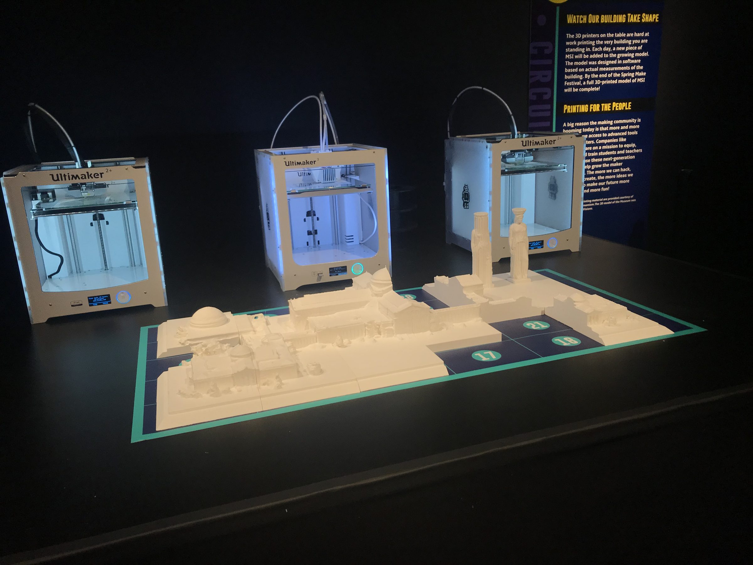 Close view of three Ultimaker 3D printers glowing on a dark table behind a mostly complete white model of the museum laid out on a teal-bordered grid.