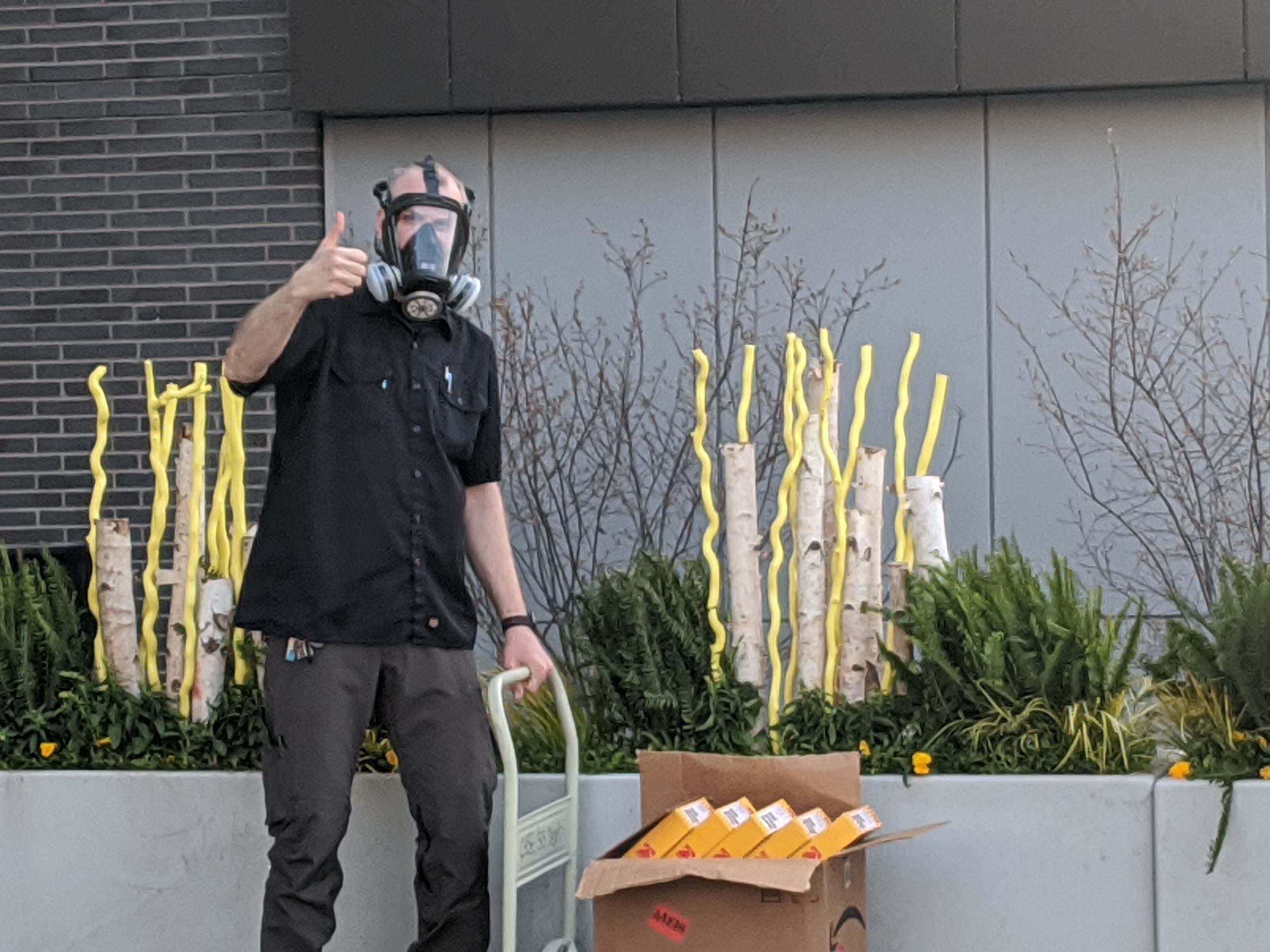 A person wearing a respirator gives a thumbs-up beside a hand truck loaded with boxes of face shields outside a building.