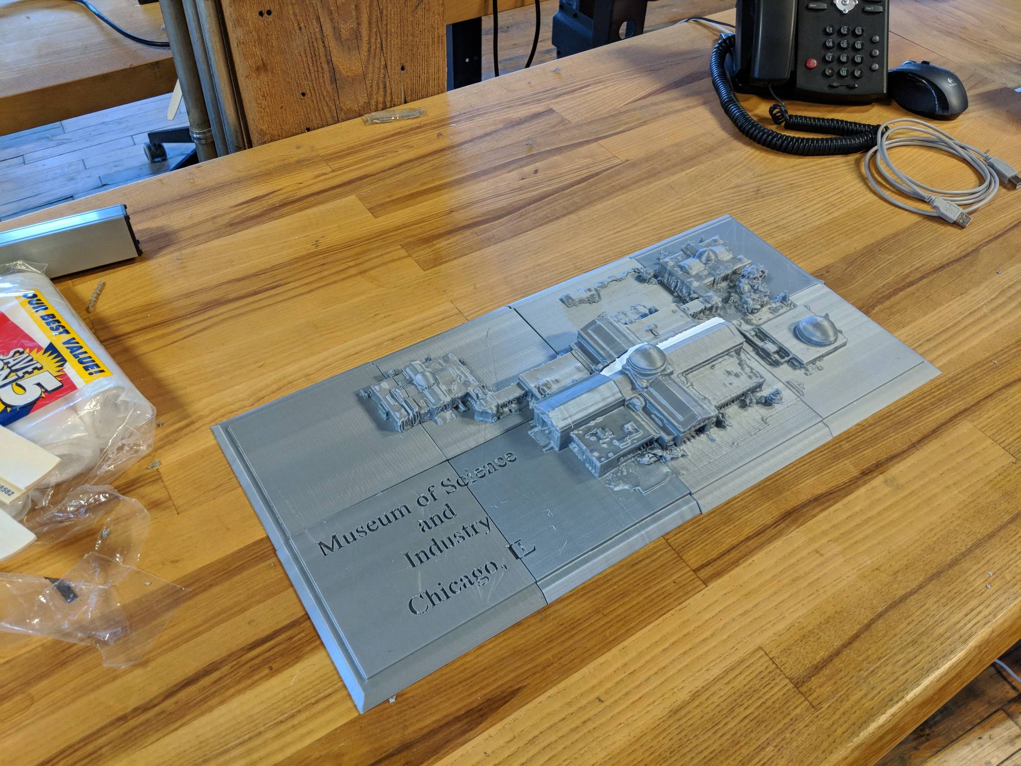 A small grey 3D-printed model of the museum on a light-blue sheet on a wooden desk.