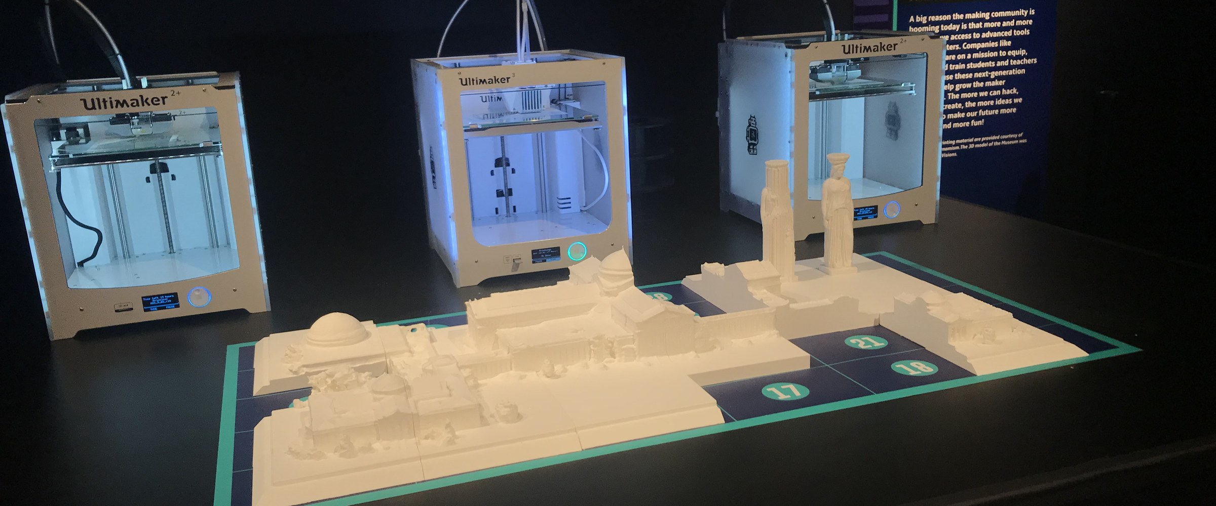 Three Ultimaker 3D printers glowing on a dark exhibit table behind a white 3D-printed model of the Museum of Science and Industry, laid out on a teal-bordered numbered grid.