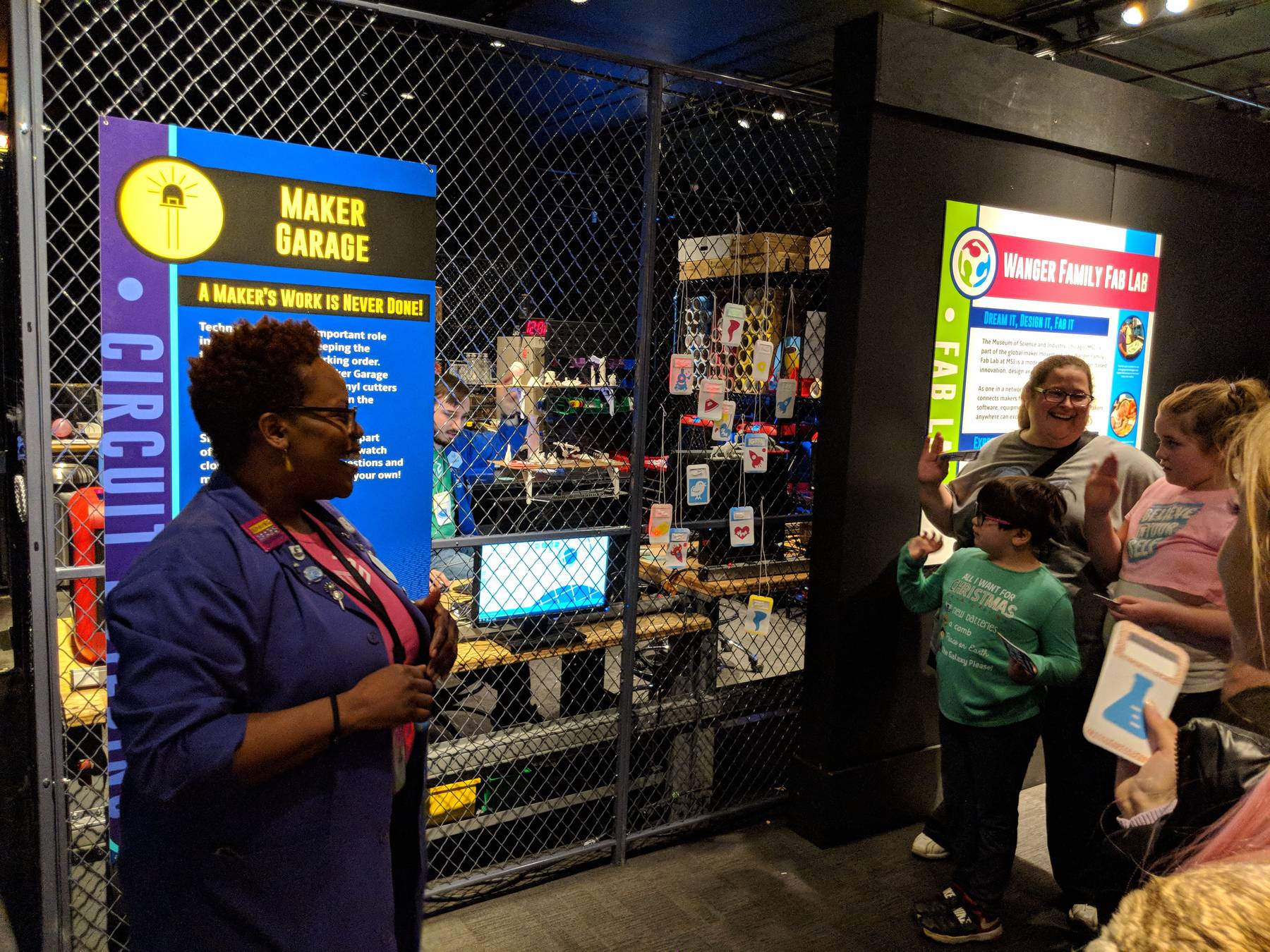 A smiling MSI staff member in front of the Maker Garage sign while children and a parent look at a display in the background.