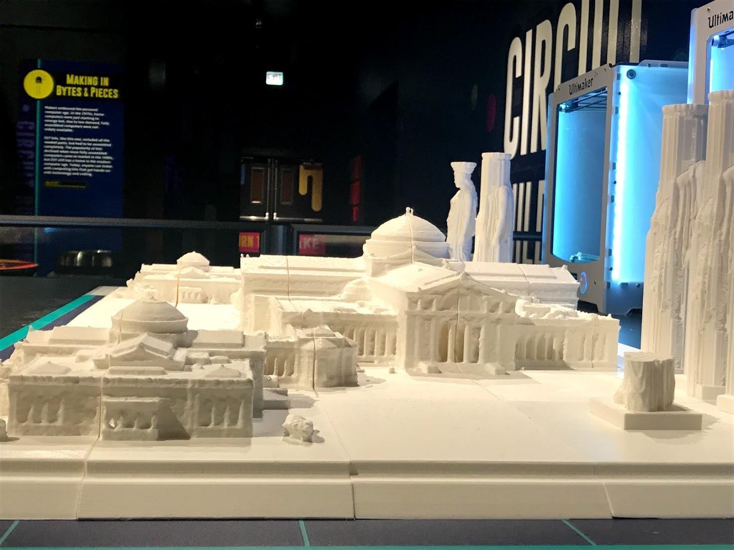 Close-up of the almost finished white 3D-printed model of the museum, with its central dome, colonnades and tall statue-topped towers, printers glowing behind.