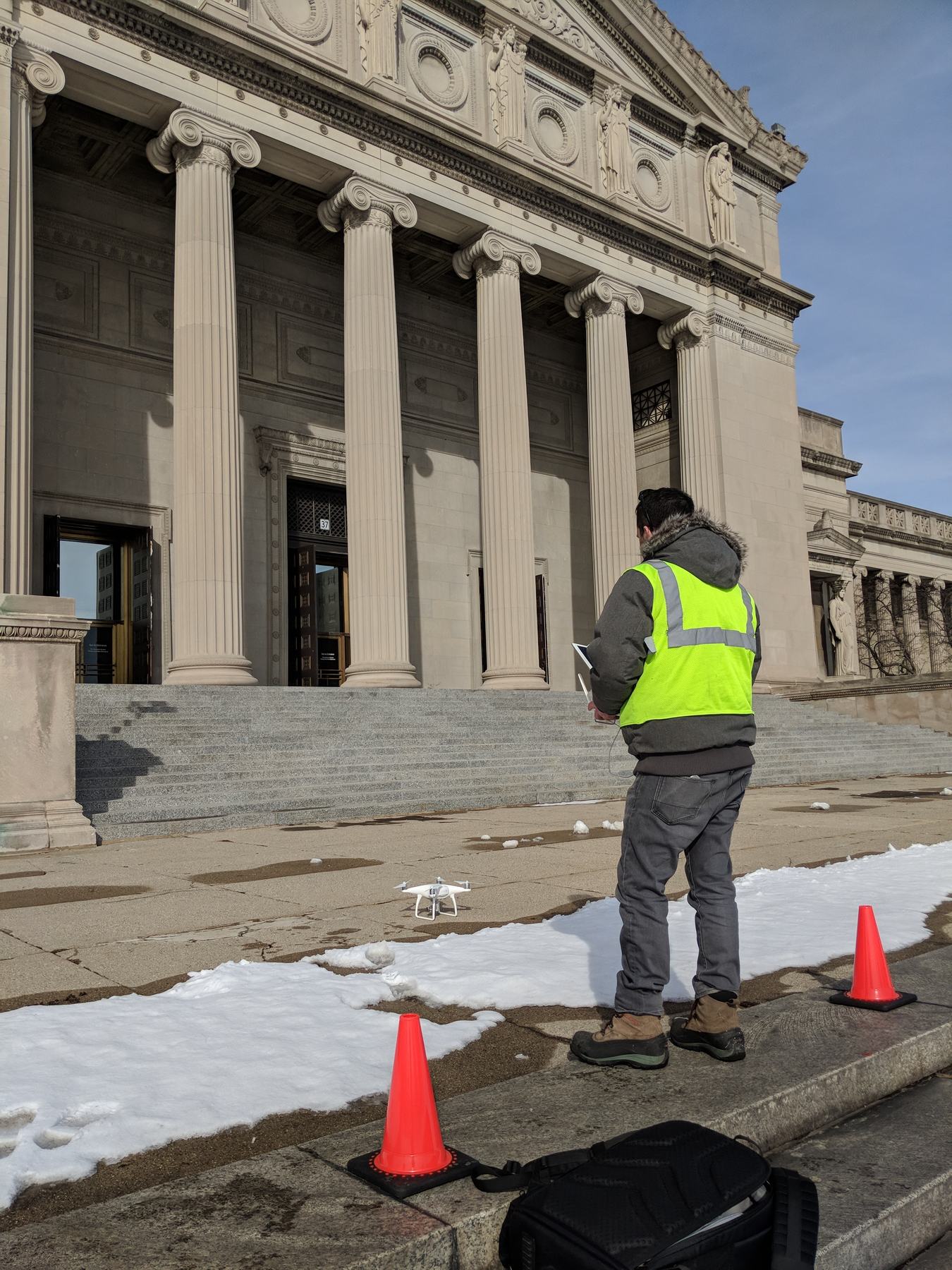 A drone pilot in a high-visibility vest stands on the museum's front steps between orange cones while a white quadcopter sits on the pavement, with the columned entrance behind.