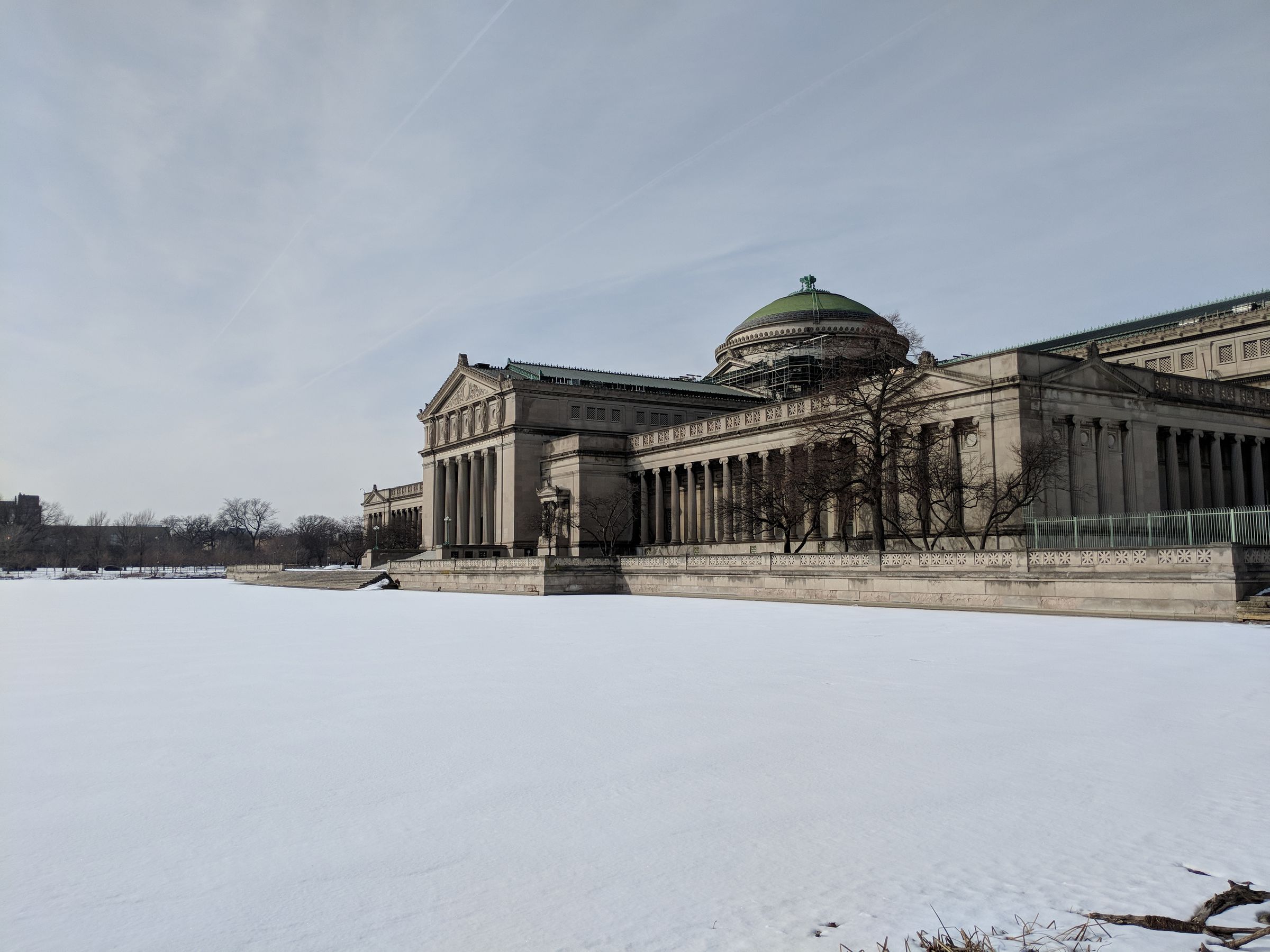 The Museum of Science and Industry's neoclassical facade and green dome seen across a snow-covered lawn under a pale sky.