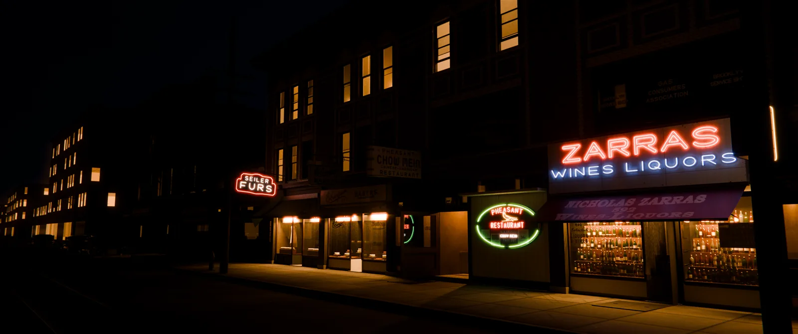 The same storefronts at night on dry pavement: the red and blue Zarras neon, the green-ringed Pheasant Restaurant neon and the Seiler Furs sign glow, the shop windows are lit, and lit windows line the buildings down the street.