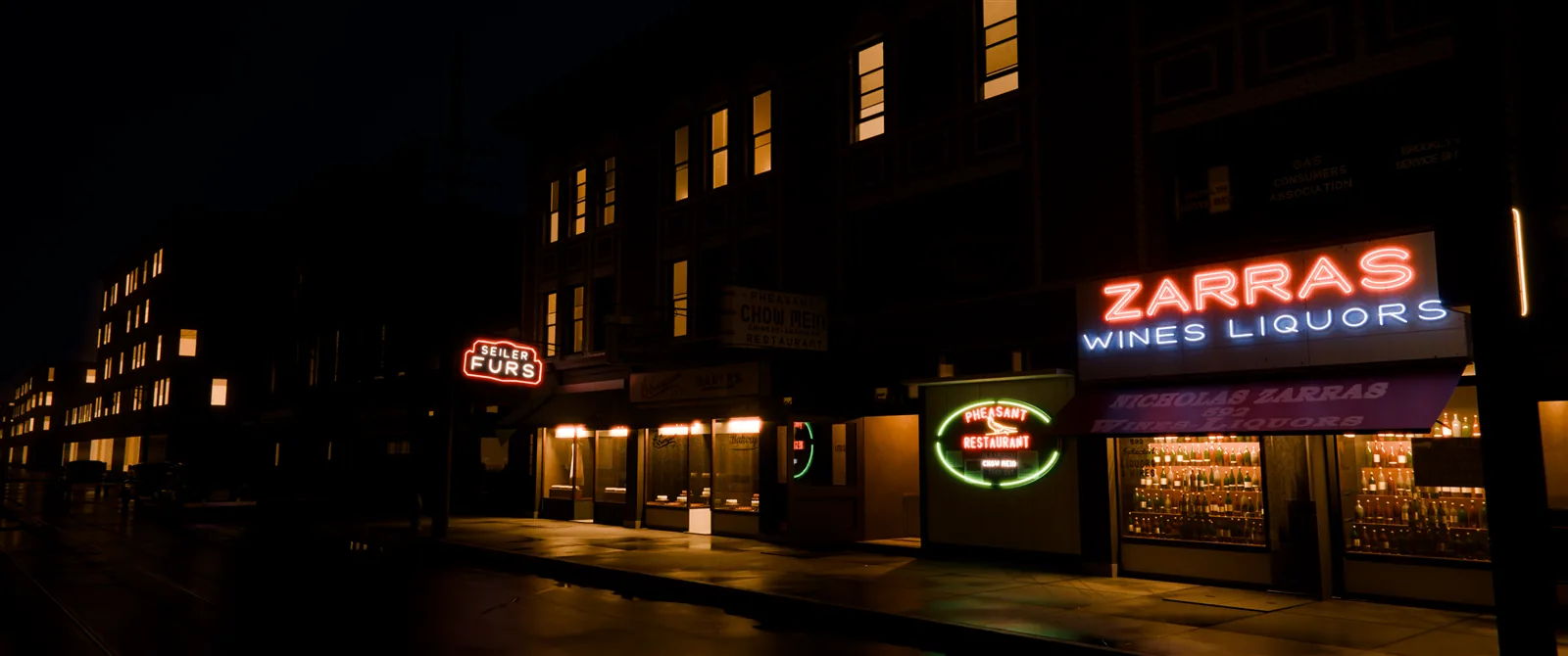 The same storefronts at night after rain: the neon signs and lit shop windows glow over a wet street and sidewalk that pick up their light.
