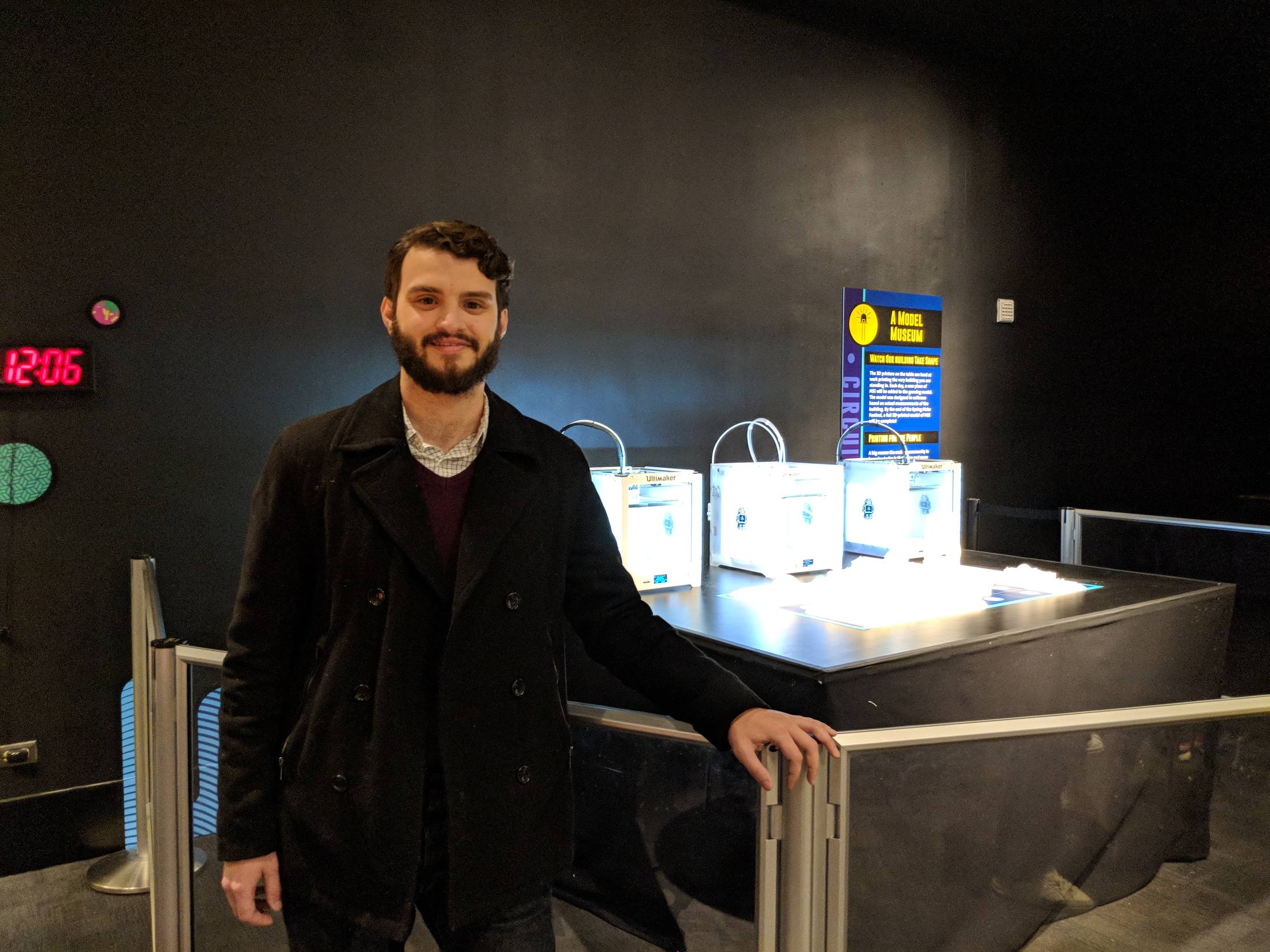 Rob Schick, smiling, in a dark coat, leaning on the railing in front of the exhibit table with its three printers and model.