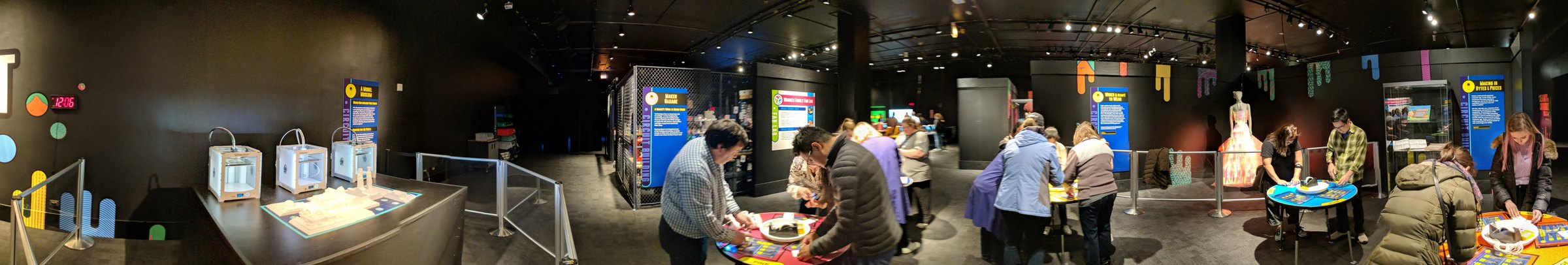 Wide panorama of the dimly lit festival hall with maker stations, visitors and the exhibit table.