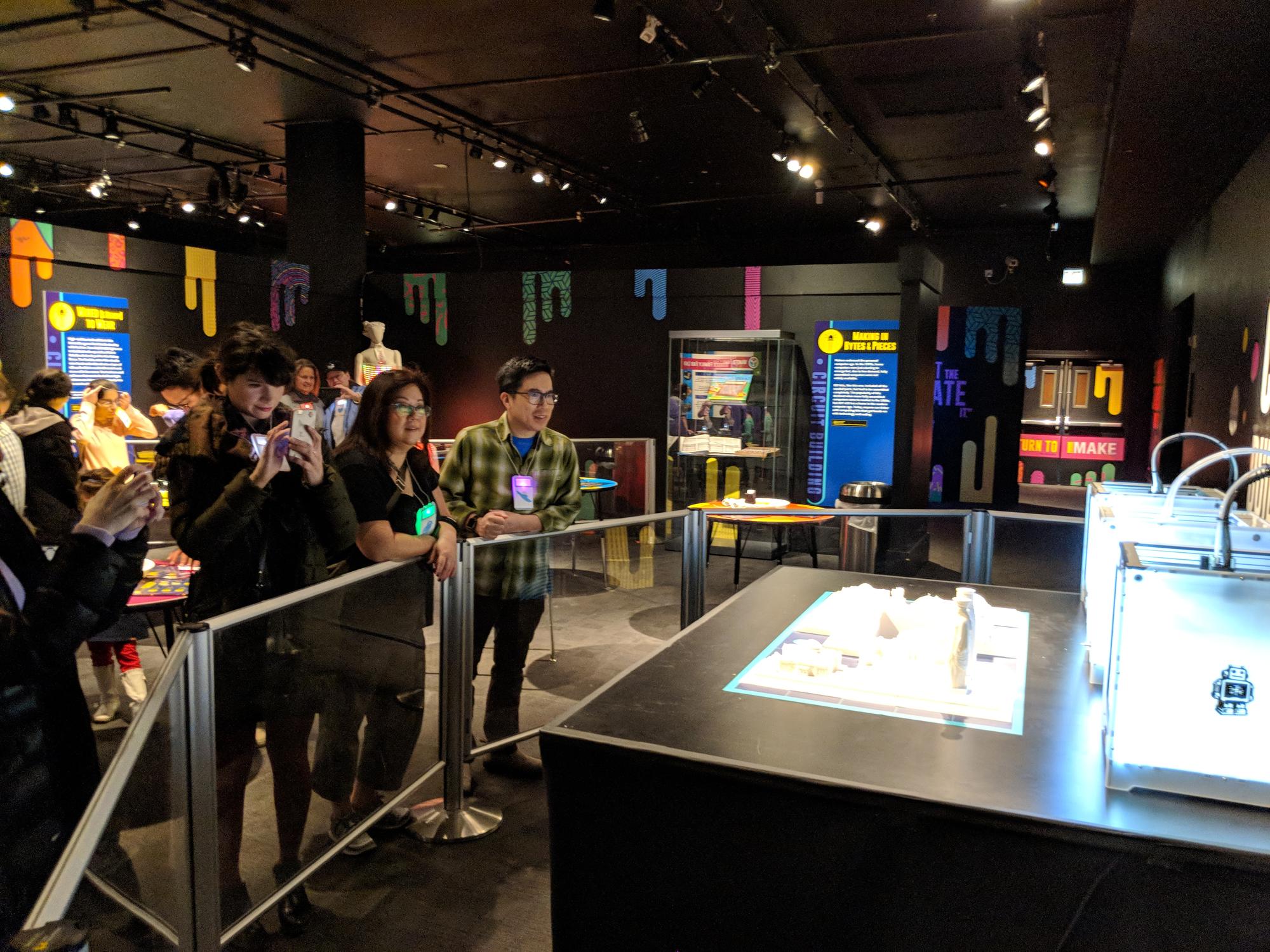 A crowd of museum visitors, including families with phones out, gathered around the exhibit table and printers.